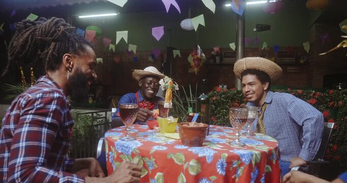 Diverse group of happy friends sitting at a decorated table during a Brazilian June festival celebration, wearing traditional straw hats and plaid shirts while talking and laughing together