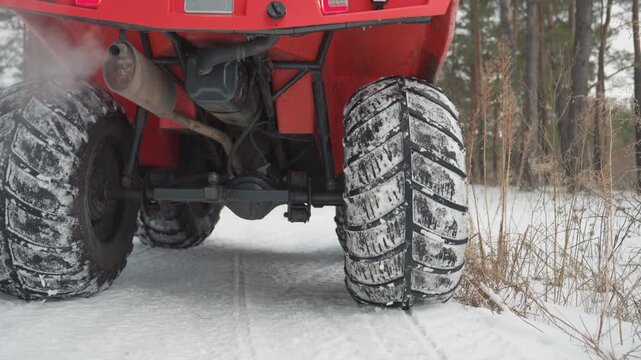 Closeup ATV tires kicking snow as red quad spins on frozen trail, rear wheel spray and visible exhaust, two riders in helmets accelerate through pine forest, hightraction offroad action