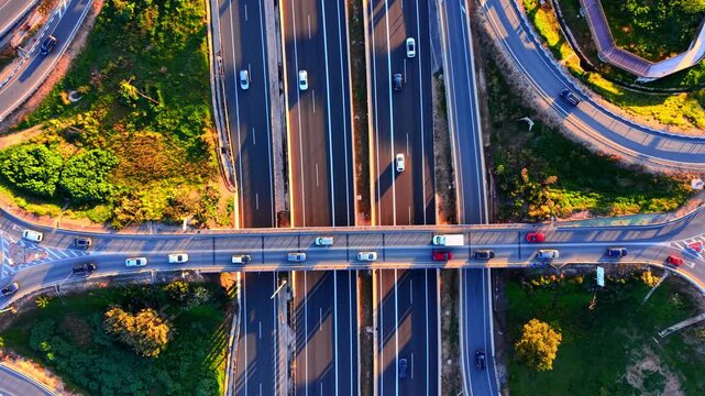 Directly above view of a bridge crossing over a multi lane highway. Several cars are visible on both levels. Surrounded by green landscape and cloverleaf ramps.