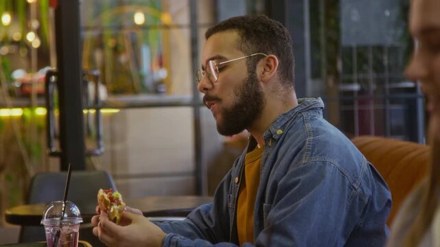 Young man with beard and glasses eating cheeseburger and having drink from straw at modern fast food restaurant