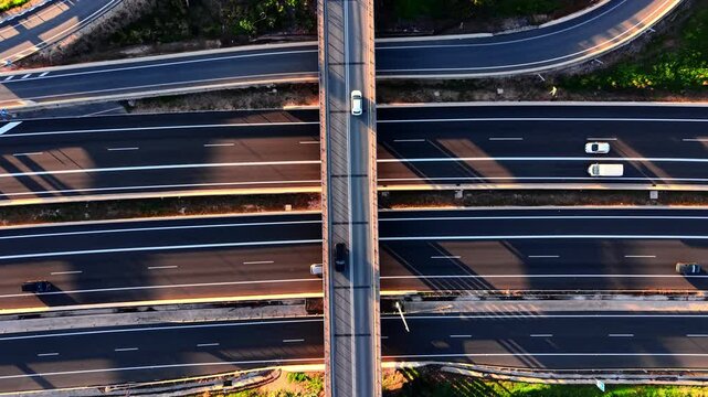 Aerial view of a road bridge crossing multiple lanes of a highway. Cars are driving in different directions. Long shadows and clean geometric patterns.