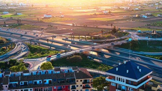 Drone perspective of complex road junction with cars driving on asphalt bridges during golden hour lighting near agricultural fields. Aerial view of multi level highway interchange at sunset.