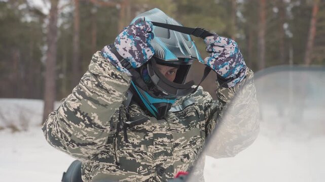 Caucasian rider adjusting helmet beside snowmobile, camo jacket and patterned gloves, mirrored goggles lowered, foggy breath in cold pine forest, methodical safety check before offroad snow ride,