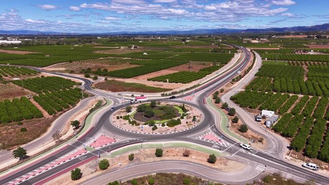 Top-down aerial view of a large modern roundabout road intersection with multiple lanes, surrounded by green citrus orchards and farmland in rural Spain under a blue sky