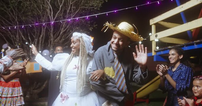 Joyful Brazilian people celebrating the traditional Festa Junina at night, featuring a couple dressed as a bride and groom in rural folk costumes dancing and waving to a cheering crowd