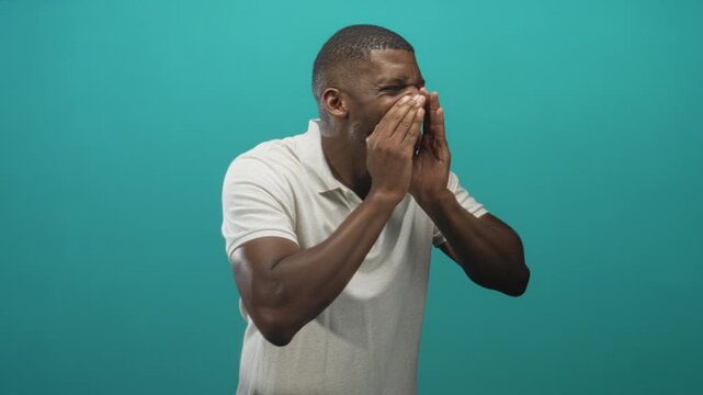 Young black man cupping hands over mouth shouting, mouth open and eyes squinting, wearing white polo against teal backdrop in studio; urgency.