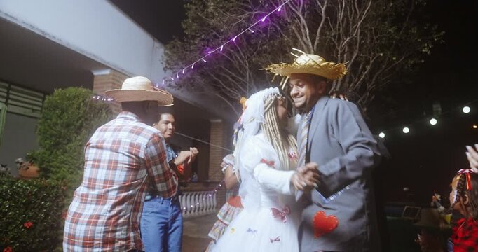 Traditional Brazilian Festa Junina celebration at night with people in caipira costumes dancing the quadrilha, featuring a bride and groom couple under festive lights and decorations