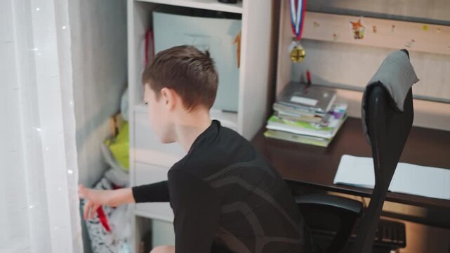 Young boy relaxing. Youth reflects near window with lights. Reflective adolescent at desk with curtains and books nearby. Young person takes break and looks out at illuminated window setting