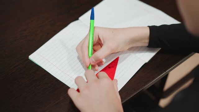 Child practices drawing. Child meticulously traces triangle using stencil on dotted grid notebook. Grandson carefully uses stencil and pen to draw triangle on dotted paper in focused manner
