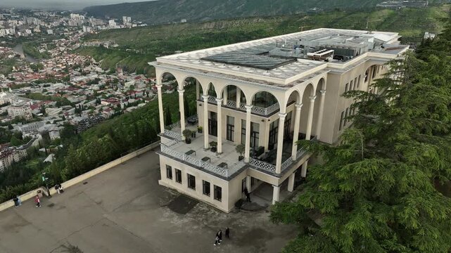 Aerial view of Mtatsminda Park funicular station and restaurant building in Tbilisi, Georgia, with the TV tower and Ferris wheel in a lush green mountain landscape.