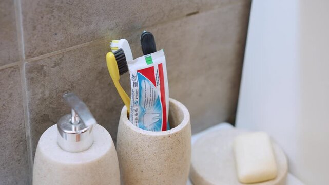 Ceramic cup filled with toothbrushes and tube of toothpaste on sink ledge, hand reaches to place green brush into holder, soap dish nearby, colorful bristles and neat arrangement, home bathroom