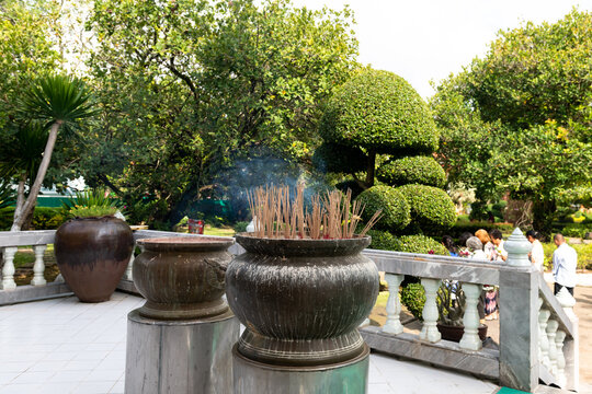 Large ceramic incense burners with burning joss sticks emitting blue smoke in a lush tropical garden of a Thai Buddhist temple, surrounded by green topiary trees and visitors under bright daylight.