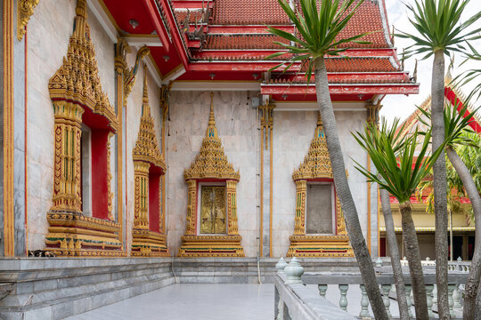 Beautifully ornate Thai Buddhist temple architecture with intricate golden window frames, marble walls, and lush green palm trees under bright daylight in Phuket.