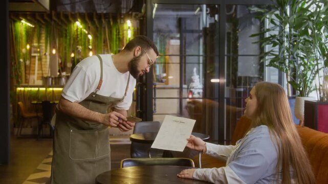 Waiter in apron taking order from female customer, pointing to her chosen dish on menu in cozy modern cafe decorated with greenery