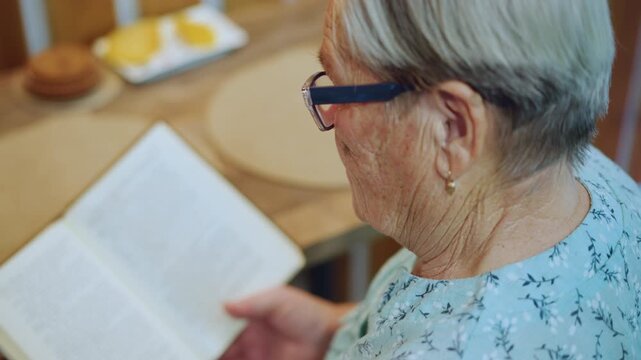 Overshoulder white grandma reading book, kitchen breakfast setup, plate and bun on table, quiet morning light, attentive expression behind glasses, pages turned slowly, peaceful domestic scene