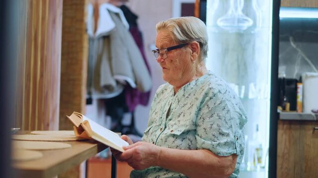 Senior lady concentrates on book details. Elderly grandmother engrossed in her reading material carefully. Senior grandmother attentively examines her book amid warm kitchen lighting ambiance