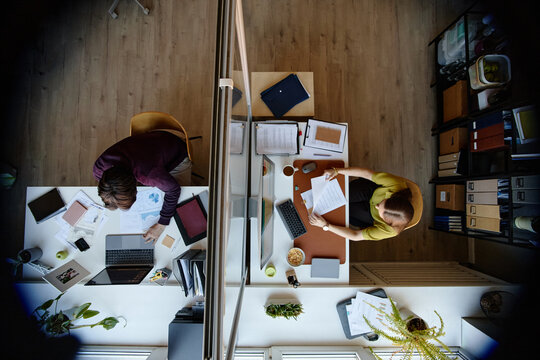 Overhead shot of young adult coworkers working at separate desks in shared office, reviewing documents and laptop data. Ideal for teamwork, productivity, office planning marketing