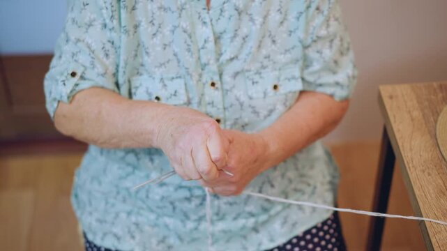 Senior lady untangling colorful thread carefully. Old woman sitting at table with yarn and flowers. Elderly lady dressed in floral attire engaging in domestic craft activity indoors