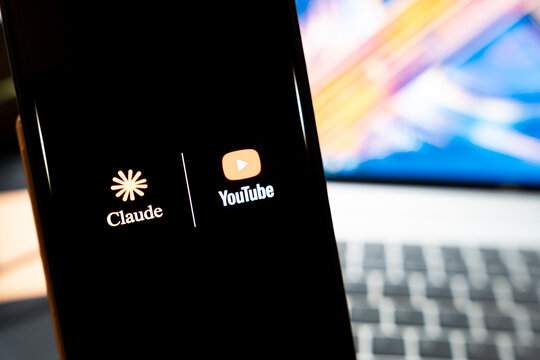 Warsaw, Poland - May 13, 2026: Smartphone displaying Claude and YouTube logos on its screen with blurred laptop keyboard in background, illustrating digital content creation and artificial intelligen