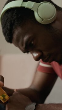 Vertical close up shot of young adult African American man in headphones looking worried while considering taking oral diabetes medication in prescription bottle at home, tilt up camera movement