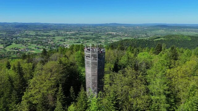 Aerial circle orbit shot of a wooden observation tower in the forest, people on the viewing platform, hills and villages in the background.