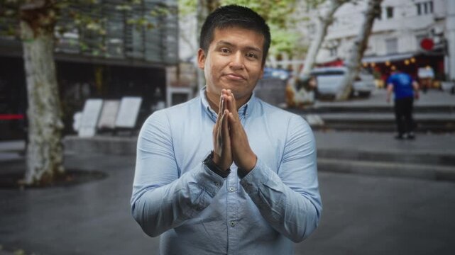 Young hispanic man wearing light blue shirt with hands pressed together in prayer, slight smile, standing on city street by steps and trees; pleading forgiveness.