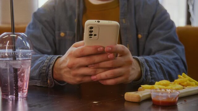 Low angle close up shot of unrecognizable man texting on his smartphone while sitting at table with fast food in restaurant