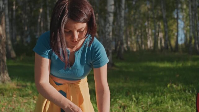 Woman chopping vegetables at campsite table, slicing yellow squash on board near bowl of mushrooms and pan, knife work closeup, sunlight and birch trees in background, intentional meal prep and rustic