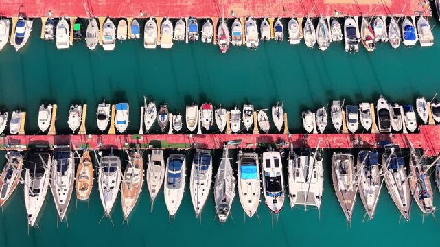 Detailed top view of white sailboats and catamarans lined up at a red floating dock in clear green water. Aerial perspective of sailboats moored at red pier.