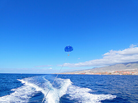 Blue parasail flying above Atlantic Ocean behind speeding boat near Tenerife coastline and mountain landscape. Sunny summer travel scene with sea waves, water sport activity and vacation adventure