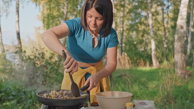 Woman cooking camping skillet by birch trees, stirring vegetable hash over portable stove, warm sunlight on green meadow, focused expression, apron and bowl on picnic setup, rustic outdoor meal prep,