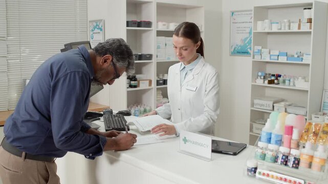 Medium shot of young woman wearing white coat as pharmacist serving adult male customer signing medication order form at checkout counter at pharmacy, copy space