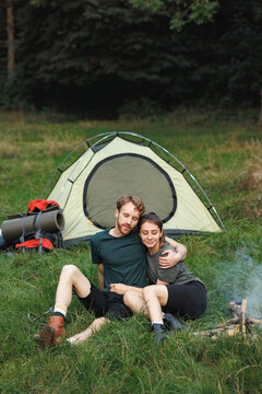 Young couple sitting near fire near tent in forest at summer