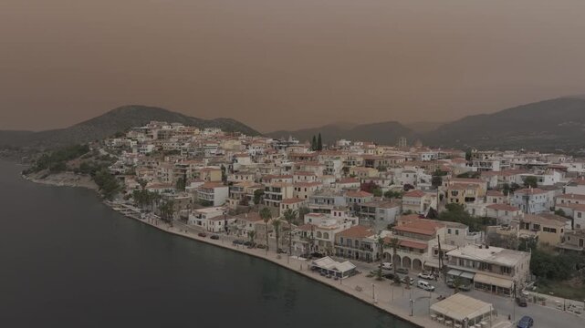 Aerial drone shot rising above Ermioni coastal town revealing expanding landscape, mountains and Mediterranean coastline under dusty orange sky