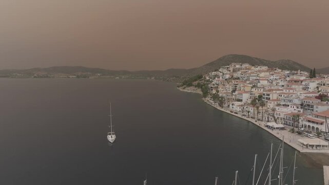 Cinematic drone pullback from sailing yacht entering Ermioni harbor in Greece, revealing the marina and coastal town under dramatic Sahara dust orange sky