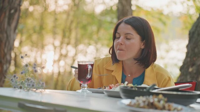 White woman camping sunset lakeside with quiet dinner, editor on creative retreat sipping wine, hands holding fork, reflective mood, shimmering water and tree silhouettes, warm glow