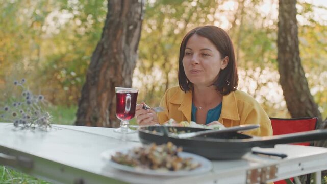 Smiling woman having cozy outdoor meal. Woman seated at campsite enjoying warm evening with drink. Content woman relishing peaceful outdoor dinner beneath golden light and tall trees