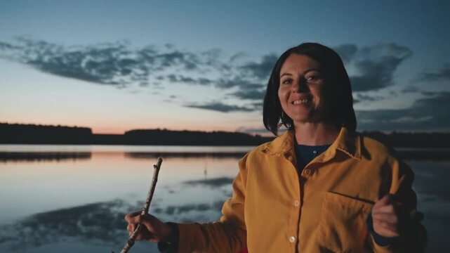 Evening lakeside camper roasting stick smiling, caucasian woman laughs while holding roasting stick, implied marshmallow moment, warm glow and ember light, storytelling energy, cozy camping vibe