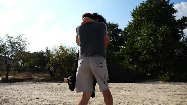 Carefree lovers having fun together on seashore at summer vacation. Man taking his beloved in air, turning around and kissing her at coastline. Happy couple spending time at sandy beach. Slow mo