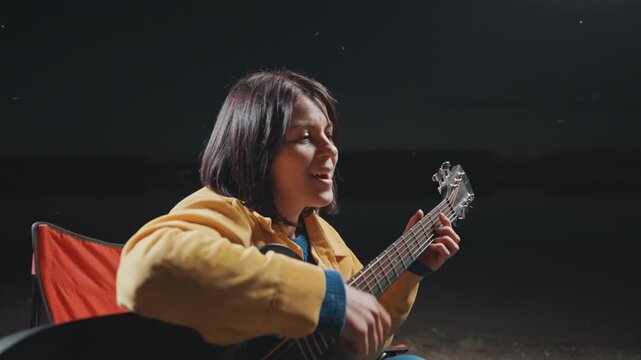 Closeup of woman singing by peaceful lake. Emotional portrait of girl softly strumming near water. Calm evening scene showing singersongwriter with acoustic instrument by tranquil lakeside