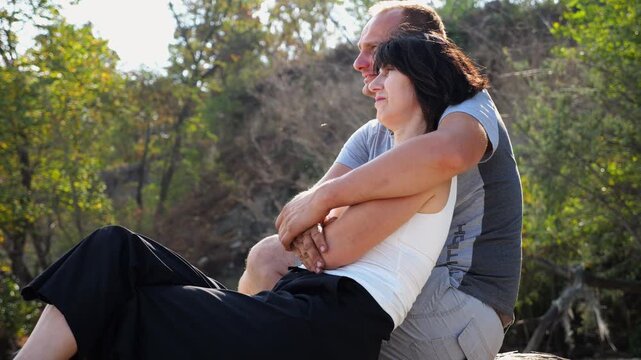 Happy pair hugs during sitting on wooden log on beach. Romantic couple admiring sea landscape at sunny day. Lovers spending time together at resort. Concept of summer vacation or holiday. Slow mo
