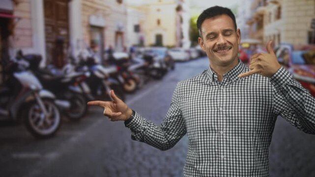 Hispanic man with moustache pointing his finger and making a phone hand gesture while smiling on a cobblestone street lined with scooters; cheerful welcome.