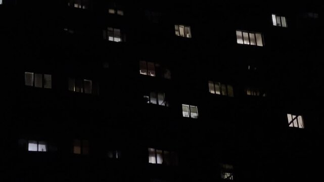 Night view on apartment building with illuminated windows. Warm lights glow from different flats, creating a quiet urban atmosphere and a sense of life behind the facades in the darkness. Slow motion
