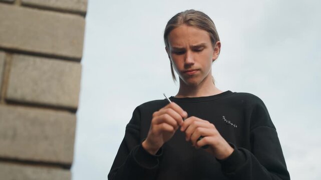 Young man prepares cigarette outdoors. Focused individual rolling cigarette beneath cloudy sky. Youthful male carefully rolling and lighting cigarette while urban environment surrounds him