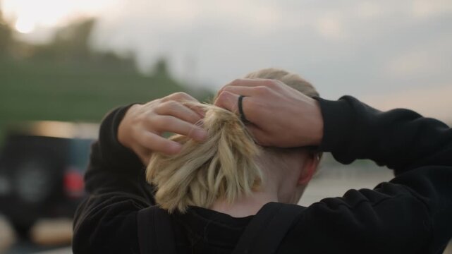 Blonde white woman back preparing run, hands gathering hair into ponytail, tightening elastic, green park and road backdrop, sunset light, hoodie, focused ritual, athletic energy, candid b roll vibe.