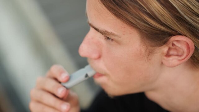 Student smokes outdoors. Young man relaxes with vape. Reflective male student pauses for thought while vaping outside. Young man in casual attire takes break while vaping on balcony