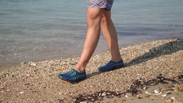 Man in water shoes walking on pebbly beach along sea on sunny day. Protective footwear preventing foot injuries while walking on sharp stones and possible sharp objects.