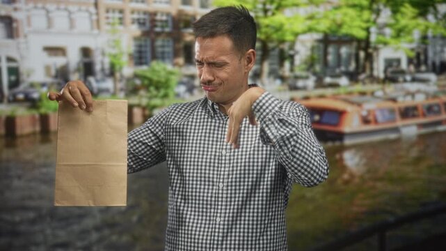 Man holding paper bag pinching nose with grimace and pointing thumb at the bag while wearing checked shirt on street by canal boat; disgust smell.