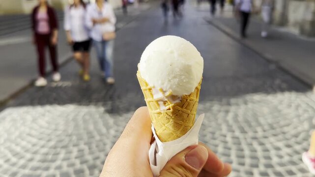POV of male hand holding an ice cream cone while walking along city street. Blurred pedestrians pass by in the background, creating a lively urban atmosphere and emphasizing the treat in arm