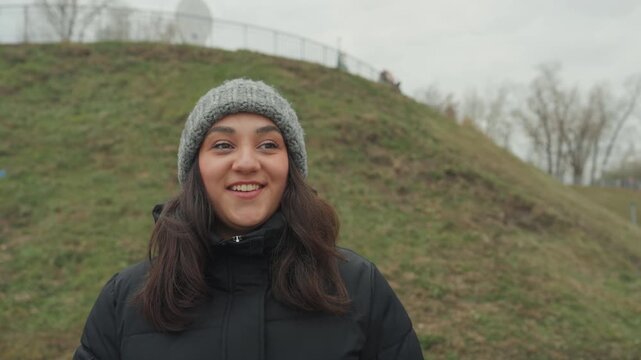 Park rhythm woman dancing clapping hands in beanie, expressive face and warm smile against grassy slope, winter coat movement shows improvisation and upbeat mood in overcast light